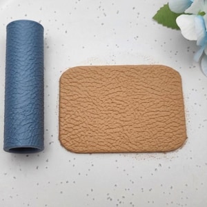 May include: A cylindrical blue object with a textured surface positioned next to a rectangular tan piece with a matching textured finish. Both items feature a consistent, pebbled grain pattern across their surfaces, displaying a matte appearance.