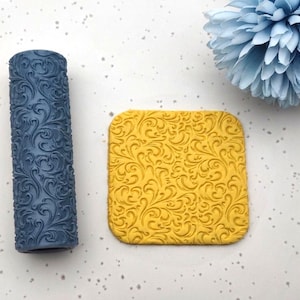 May include: A dark blue patterned rolling pin and a yellow square with a matching design. The floral pattern is embossed on both items. A light blue flower is in the background.