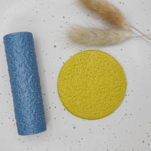 May include: A textured, cylindrical blue object and a yellow, circular object are displayed on a speckled white surface. Dried, tan-coloured grass-like stems are also present. The objects are likely decorative or functional items.