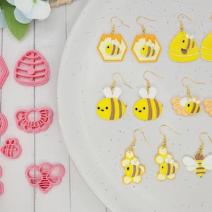 May include: A selection of pink bee-shaped cookie cutters and yellow bee earrings with gold-coloured hooks. The earrings showcase diverse designs, including honeycombs and bees with white wings. The items are displayed on a white surface.