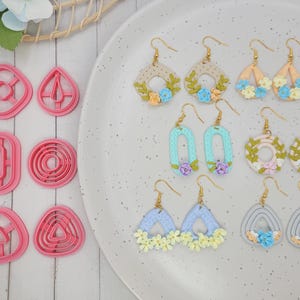 Può includere: Orecchini fatti a mano in varie forme e colori, tra cui beige, blu e pesca, adornati con disegni floreali. Sono visibili anche taglierini in argilla polimerica rosa a forma geometrica.