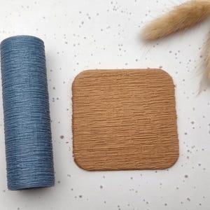 May include: A blue cylindrical object with a textured surface and a square, brown coaster with rounded corners. Two dried, fluffy, tan-coloured decorative stems are also present.
