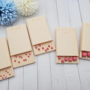 Peut inclure: Cinq plateaux de tri de perles rectangulaires en plastique beige avec couvercles coulissants. Chaque plateau est marqué avec des tailles en millimètres, notamment 2 mm, 3 mm, 4 mm, 5 mm, 6 mm, 7 mm, 8 mm, 9 mm, 10 mm, 12 mm et 15 mm, contenant de petites perles sphériques roses.