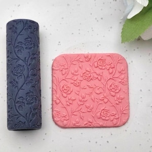 May include: A dark blue cylindrical rolling pin and a square pink slab, both featuring an embossed rose and vine pattern. The cylindrical tool is positioned vertically next to the flat square piece, which displays the same floral texture.