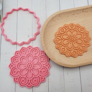 Op de afbeelding: Een roze plastic koekjesvorm met een gekartelde buitenrand, een roze plastic stempel met een gedetailleerd bloemenmandala-patroon en een oranje geperste kleischijf met hetzelfde complexe bloemendesign als de stempel.