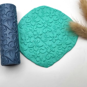 May include: A dark blue floral patterned rolling pin and a teal clay sheet with a matching floral design. The rolling pin is cylindrical, and the clay sheet is a rounded shape. Dried ornamental grass is in the background.