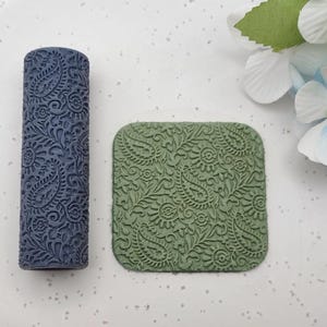 May include: A dark blue cylindrical patterned roller and a square sage green tile featuring an identical embossed paisley and floral design. The roller has a textured surface for creating impressions, while the tile displays the finished pattern.