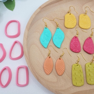 Könnte beinhalten: Eine Sammlung handgefertigter Ohrringe in verschiedenen Farben und Formen, darunter Blatt-, Oval- und Rechteckdesigns. Die Ohrringe werden auf einem Holztablett zusammen mit rosa Ohrringausstechern präsentiert. Die Ohrringe weisen strukturierte Muster und goldfarbene Haken auf.