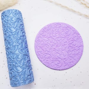 May include: A blue rolling pin and a purple circular stamp, both with an embossed floral design. The rolling pin is cylindrical, and the stamp is round. The items are likely for baking or crafting.