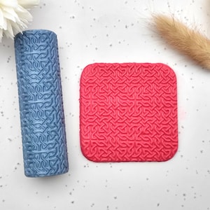 May include: A blue cylindrical item and a red square item, both featuring a repeating Celtic knot design. They are set on a white surface, with a white flower and dried grass sprigs in the background.