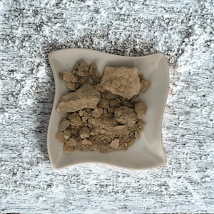 Peut inclure: Un petit plat blanc cassé aux bords ondulés rempli de morceaux et de granulés d'une substance brun clair. Le plat est posé sur une surface texturée blanche et grise.