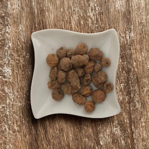 May include: A white, square-shaped dish filled with a pile of brown, textured tiger nuts. The dish sits on a wooden surface with a natural grain pattern. The nuts are various sizes and shapes.