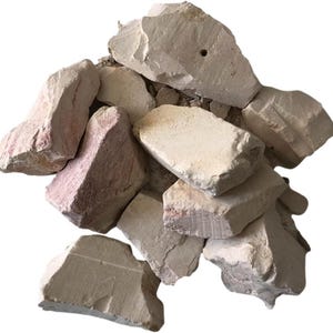 Puede incluir: Un montón de trozos de arcilla cruda, de varios tamaños y formas. La arcilla es principalmente de color beige claro, con algunas piezas que muestran tonos rosados-rojizos. La textura rugosa y las formas irregulares sugieren un material natural sin procesar.