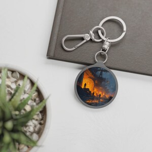 May include: A gray keychain with a round pendant featuring an artistic depiction of a fiery industrial scene. The keychain has a silver clasp and a key ring. The background includes a succulent plant in a white pot and a gray notebook.