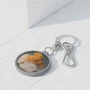 May include: A round, gray keychain with a silver clasp. The keychain features a circular image of a landscape with orange clouds and a structure. The keychain is on a white surface.