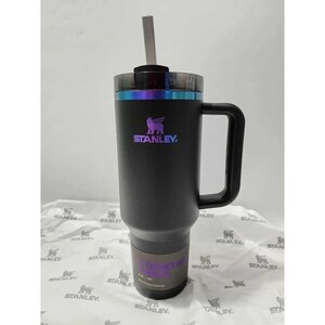 Puede incluir: Un vaso Stanley negro con asa y pajita. El vaso tiene el logotipo de Stanley en colores iridiscentes. El vaso est&aacute; sobre una superficie blanca con un patr&oacute;n repetido del logotipo de Stanley.