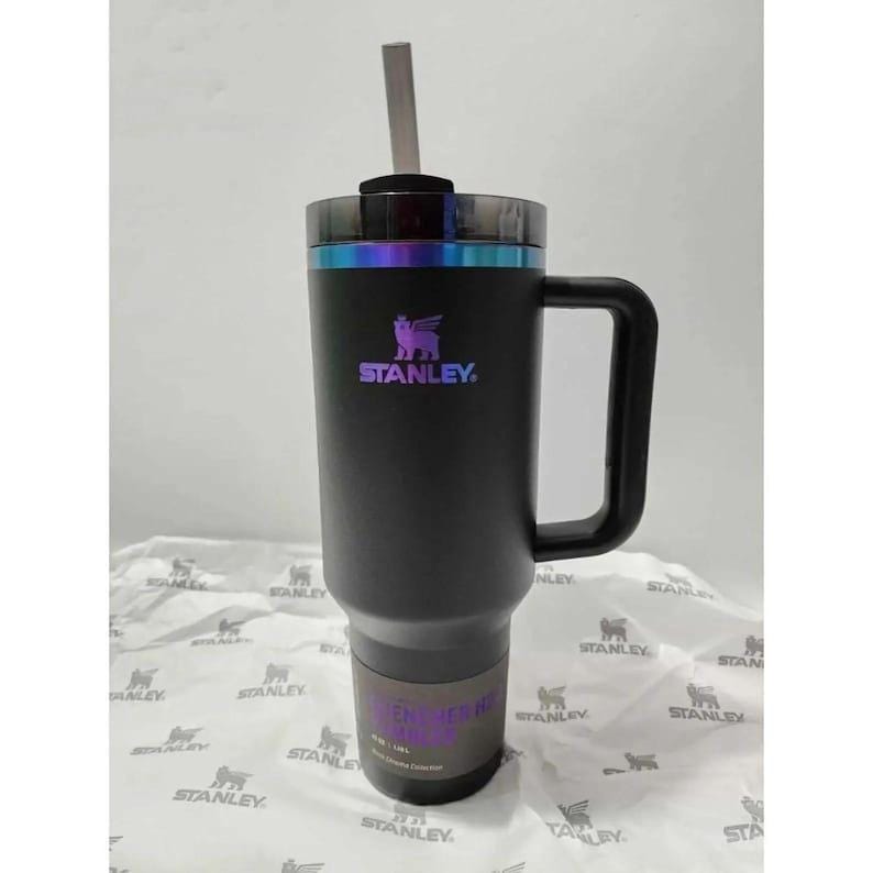 Puede incluir: Un vaso Stanley negro con asa y pajita. El vaso tiene el logotipo de Stanley en colores iridiscentes. El vaso est&aacute; sobre una superficie blanca con un patr&oacute;n repetido del logotipo de Stanley.