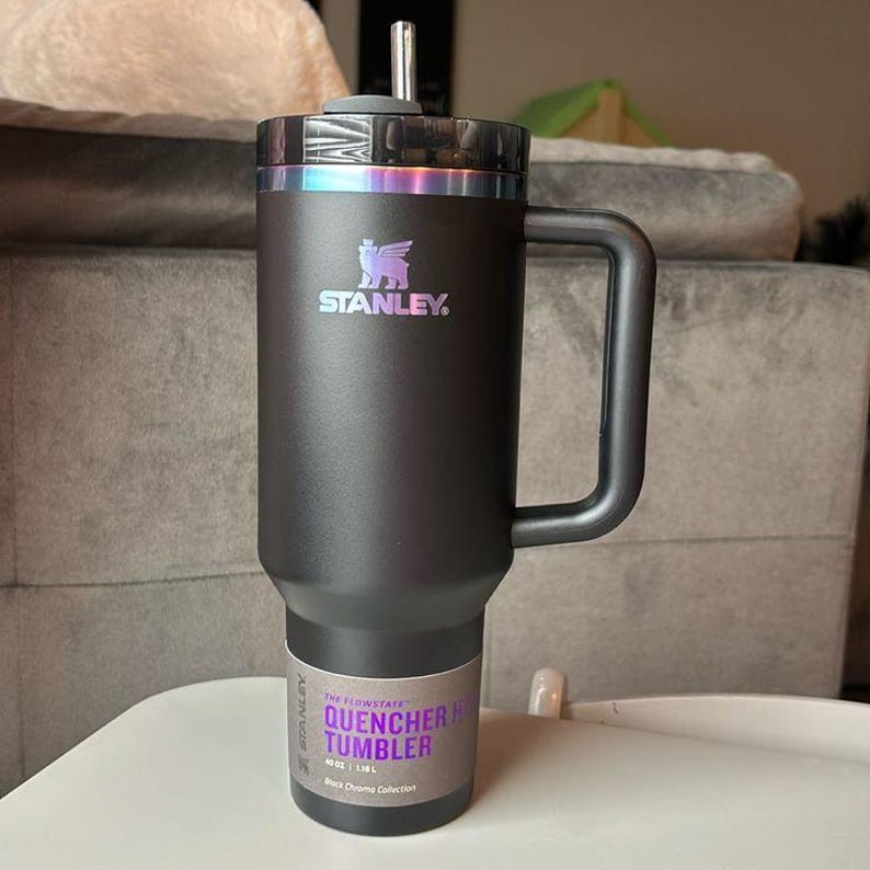Puede incluir: Un vaso Stanley Quencher H2.0 Flowstate negro con asa y pajita. El vaso tiene una tapa negra con una banda iridiscente hologr&aacute;fica. El logotipo de Stanley es visible en la parte delantera. El vaso es de 1,18L.
