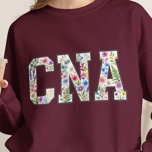以下が含まれることがあります： ブロックフォントで「CNA」の文字がプリントされたマルーンカラーのクルーネックトレーナー。文字の中は、白い背景に様々な野花、緑の葉、小さな花の小枝が描かれたカラフルなボタニカル柄で埋め尽くされています。