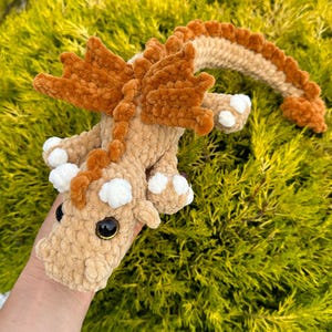 May include: A crocheted dragon toy in tan and brown, featuring orange wings and spine. The dragon has white spots on its feet and black eyes. The soft toy is set against a backdrop of green foliage.