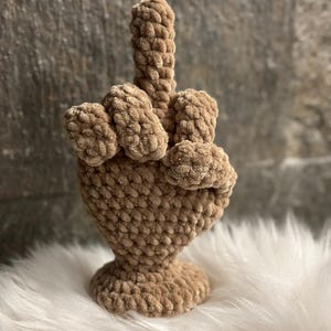 Op de afbeelding: Een beige gehaakte sculptuur van een hand die de middelvinger opsteekt. De sculptuur is gemaakt van zacht, getextureerd garen. Het staat op een witte, pluizige ondergrond. De basis is rond en de vingers zijn duidelijk gedefinieerd. Een uniek decoratief item.