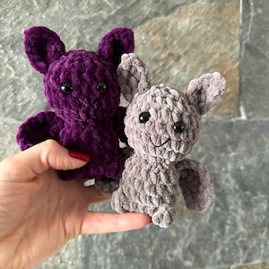 May include: Two plush crochet bats. One is a deep purple, the other is gray. Both have black button eyes and small wings. The bats are handmade and have a soft, textured appearance. They are likely intended as toys or decorative items.