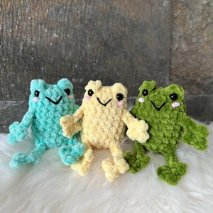 May include: Three crocheted frog toys in teal, yellow, and green. Each frog has a smiling face with black eyes and pink cheeks. The frogs are displayed on a white, fluffy surface.