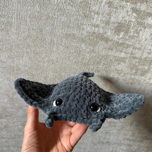May include: A handmade, plush gray manta ray with black eyes and a small trunk. The soft toy has large, wing-like fins and is held in a person's hand. The manta ray is made of a fuzzy material.