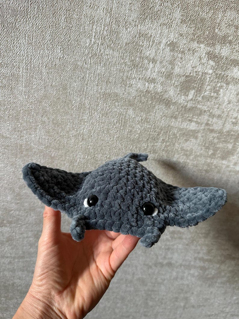 May include: A handmade, plush gray manta ray with black eyes and a small trunk. The soft toy has large, wing-like fins and is held in a person's hand. The manta ray is made of a fuzzy material.