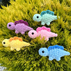 May include: Six crocheted dinosaur toys in various colours, including yellow, pink, purple, blue, and green. Each dinosaur has a different colour scheme and black button eyes. The toys have a textured, plush appearance.