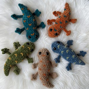 May include: Five plush lizard toys made of soft velvet yarn. Each lizard features a distinct color: teal, orange, olive green, tan, and blue. They have embroidered V-shaped patterns on their backs and large, round, black plastic safety eyes.