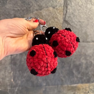 Pode incluir: Três chaveiros de joaninha vermelhos e pretos. As joaninhas são feitas de um material texturizado, com manchas pretas e uma cabeça preta. Os chaveiros são presos a uma corrente prateada.