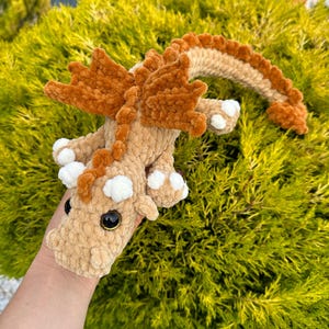 May include: A crocheted plush dragon toy with brown and white fur. The dragon has a long tail, large wings, and a friendly face with big eyes.