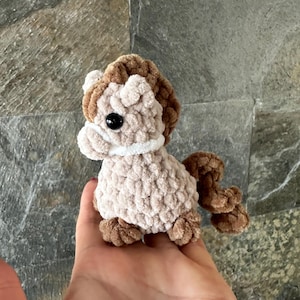 May include: A small, handmade crochet horse toy in shades of tan and brown. The plush toy features a fluffy mane, tail, and hooves, with a white collar and black eyes. The horse is designed to be a soft, cuddly toy.