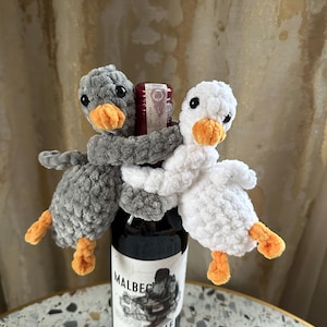 Może przedstawiać: Butelka wina Malbec jest ozdobiona dwoma szydełkowymi przytulankami do butelek wina w kształcie gęsi. Jedna gęś jest szara, druga biała, obie z pomarańczowymi dziobami i stopami. Butelka stoi na nakrapianym stole.