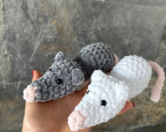 May include: Three crocheted toy rats in a hand. One is gray, one is white, and one is gray. Each has a pink nose and ears, black eyes, and a pink tail. The toys are made of a soft, textured yarn.