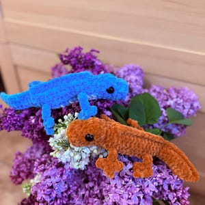 May include: Two plush, crocheted lizards rest on a bed of purple and white lilacs. One lizard is bright blue, the other is orange. Both have black eyes and are posed as if crawling. The background is a light wooden surface.