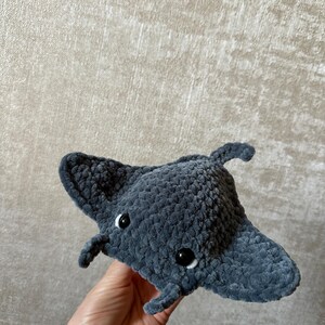 May include: A handmade, plush, gray manta ray toy with black button eyes. The soft, crocheted toy has a rounded body and two wing-like fins extending from the sides. The manta ray has two small, rounded appendages near the front.