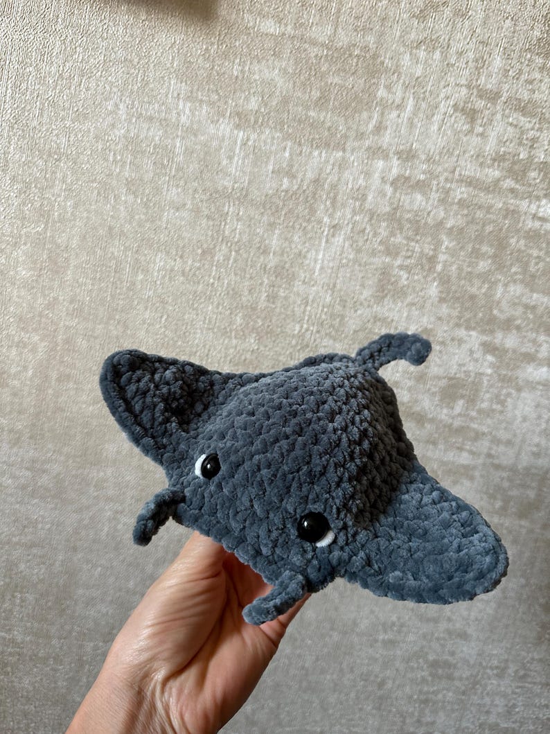 May include: A handmade, plush, gray manta ray toy with black button eyes. The soft, crocheted toy has a rounded body and two wing-like fins extending from the sides. The manta ray has two small, rounded appendages near the front.
