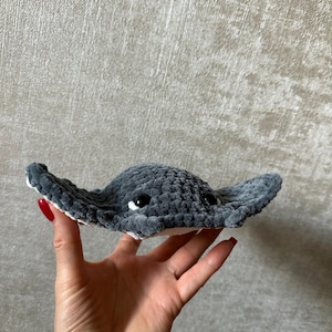 May include: A handmade, plush gray stingray toy with white accents and black eyes. The soft, crocheted toy is held in a hand, showcasing its detailed craftsmanship and cuddly appearance. The toy's wings are spread wide.