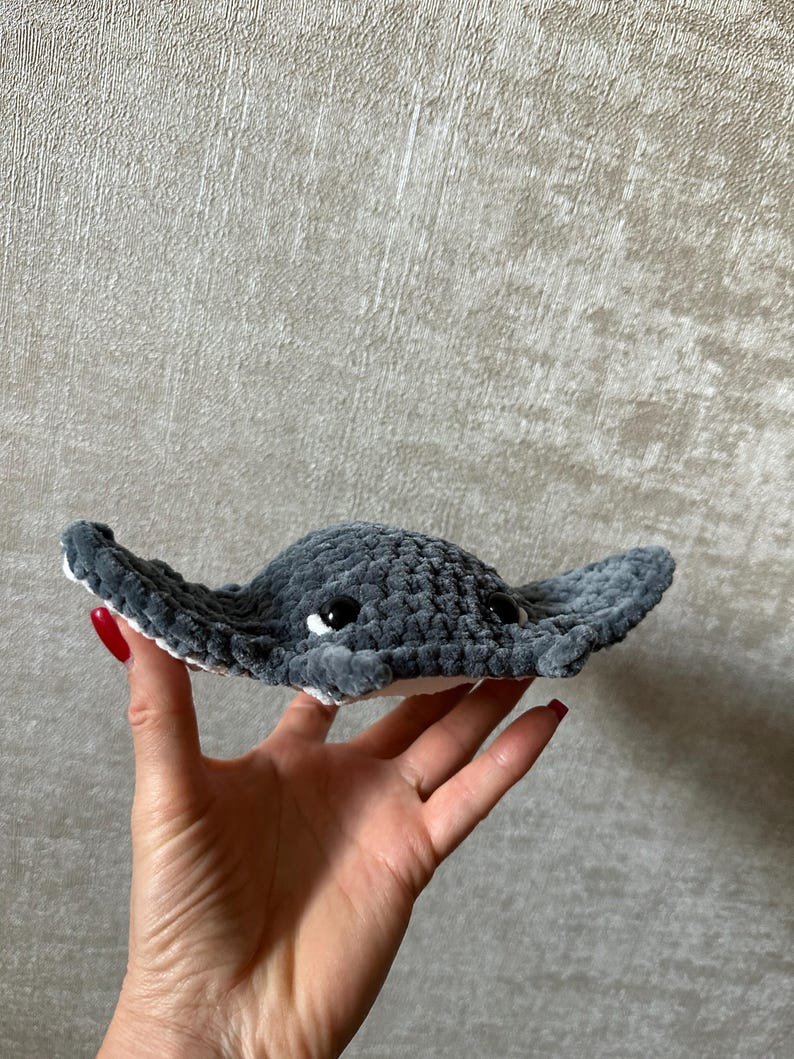 May include: A handmade, plush gray stingray toy with white accents and black eyes. The soft, crocheted toy is held in a hand, showcasing its detailed craftsmanship and cuddly appearance. The toy's wings are spread wide.
