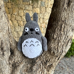 May include: A gray and white crocheted Totoro plush toy sitting on a tree branch. The plush toy has large eyes and a white belly with a pattern of triangles.