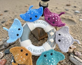 May include: A collection of handmade crochet stingrays in various colors, including blue, purple, and yellow, arranged around a life preserver that says "WELCOME ON BOARD". The scene is set on a sandy beach with seashells.