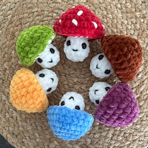 May include: A selection of crocheted mushroom figures in various colours. The mushrooms are red with white spots, green, orange, brown, purple, and blue. Each mushroom has a white stem with a smiling face and black eyes, arranged on a woven mat.