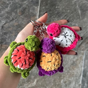 Puede incluir: Tres llaveros de tortuga de ganchillo de felpa con caparazones inspirados en frutas. Una tortuga tiene un caparazón de sandía verde y rojo, otra uno morado y naranja, y la tercera uno de fruta del dragón rosa y blanco con semillas negras.