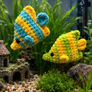 May include: Two handmade, plush fish toys in an aquarium. One fish is yellow and blue, the other is yellow and green. The fish have black eyes and are surrounded by aquatic plants, gravel, and a miniature stone castle.