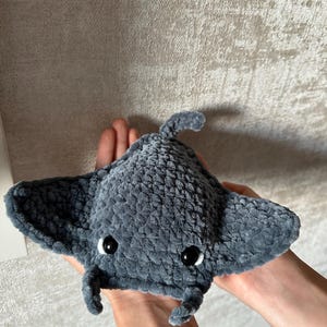 May include: A handmade, plush gray manta ray toy with black button eyes. The soft, textured fabric creates a cuddly appearance. The toy features a rounded body, wing-like fins, and a small tail, perfect for a child's playtime or nursery decor.