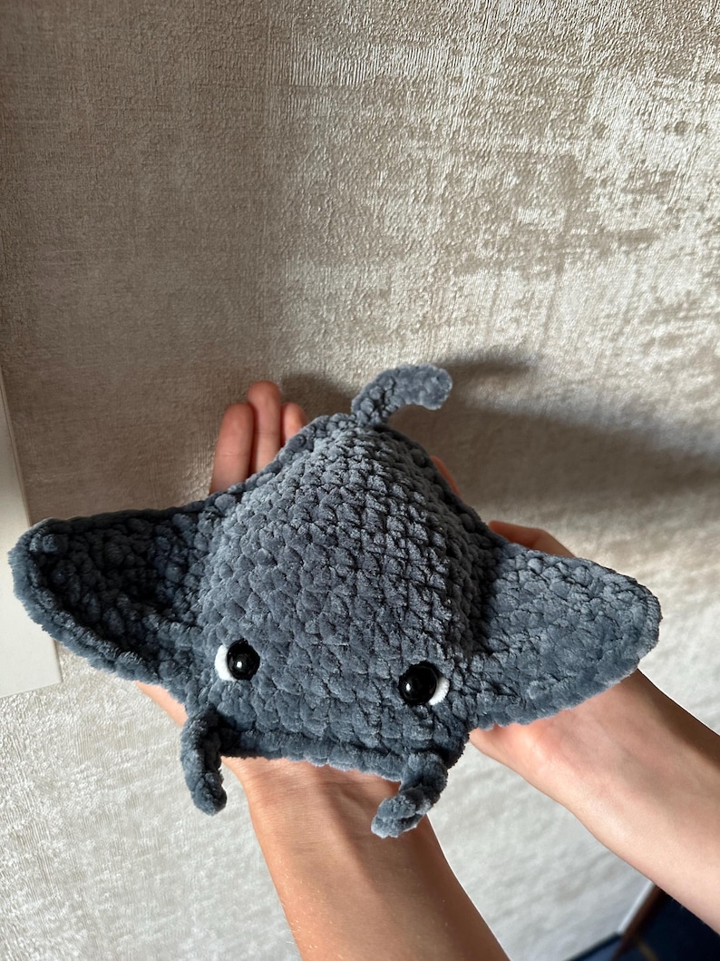 May include: A handmade, plush gray manta ray toy with black button eyes. The soft, textured fabric creates a cuddly appearance. The toy features a rounded body, wing-like fins, and a small tail, perfect for a child's playtime or nursery decor.