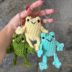 May include: Three crochet frog keychains in yellow, green, and blue. Each frog has black eyes, a smiling mouth, and pink cheeks. The keychains are attached to silver metal rings.