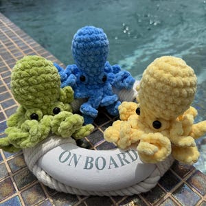 May include: Three crocheted octopus toys in shades of green, blue, and yellow, resting on a white life preserver. The life preserver has the words "ON BOARD" printed on it. The toys are placed near a pool.